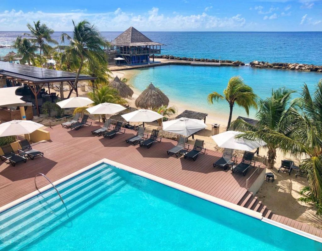 Piscina ao ar livre com espreguiçadeiras e guarda-sóis, mais vista do mar pertinho. Em hotéis em Curaçao.